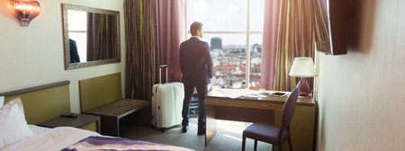 Young businessman with luggage in hotel room. Successful man in suit looking at window, arriving to businesstrip or vacation, copy spaceの写真素材
