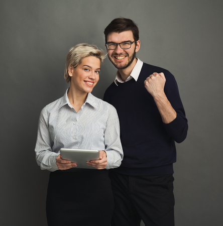 Happy couple in formal clothes posing with digital tablet, celebrating success, gray studio background, copy spaceの写真素材