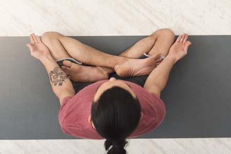Young man practicing yoga, meditation exercises. Guy does lotus pose for relaxation, sitting on mat at gym. Healthy lifestyle, copy spaceの写真素材