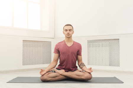 Young man practicing yoga, meditation exercises. Guy does lotus pose for relaxation, sitting on mat at gym. Healthy lifestyle, copy spaceの写真素材