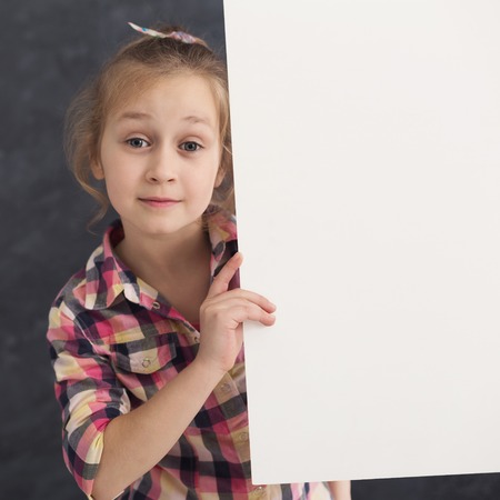 Happy little girl holding white blank banner. Excited child showing paper sheet for sales advertisement, copy space. Commercial ad mockupの写真素材