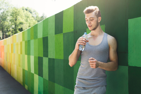 Man runner is having break, drinking water while jogging in city, leaning at green painted wall, copy spaceの写真素材