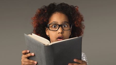 Surprised little adrican-american girl in glasses reading book on gray studio background, copy space. Back to school conceptの写真素材