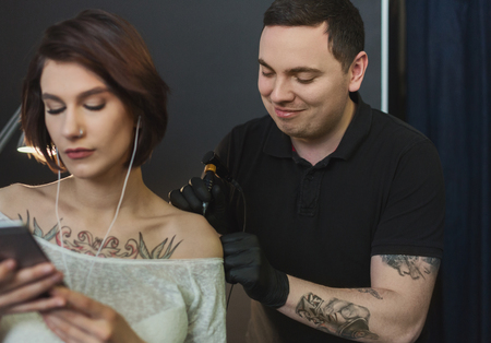 Professional tattooer doing picture on client hand by machine while girl listening music and texting on smartphone. Tattoo art on body. Popular body modification, modern lifestyle, copy spaceの写真素材