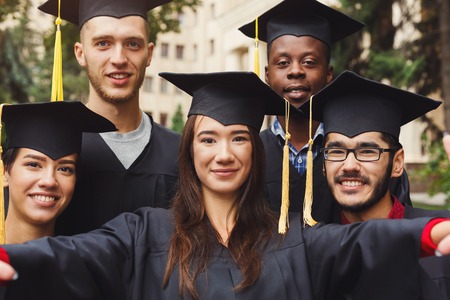 Multiethnic students taking group selfie on smartphone to celebrate their graduation. Education, qualification and gown concept.の写真素材