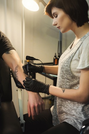 Concentrated woman tattooist doing tattoo with tattoo machine on male arm. Unusual female profession, copy spaceの写真素材