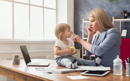 Happy mother working at office with her baby. Young woman talking on phone while spending time with her child. Business, motherhood, multitasking and family concept.の写真素材