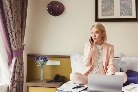 Portrait of serious businesswoman sitting on bed, using laptop and talking on smartphone. Woman working in hotel room. Business lady went to business trip and stayed at the hotel, copy spaceの写真素材