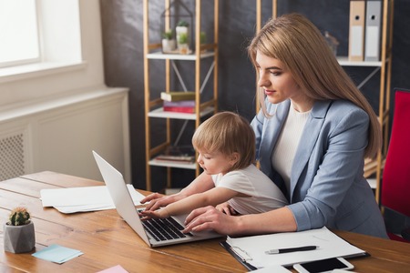 Beautiful business mom working on laptop in office while spending time with her cute baby. Business, motherhood, multitasking and family concept.の写真素材