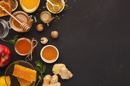 Honey assortment on black table. Top view on full glass jars, honeycombs, pollen bowl, nuts, ginger,...