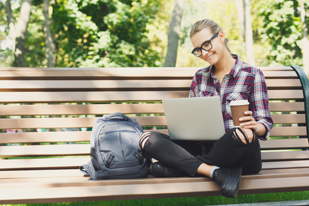 Young happy woman using laptop and drinking coffee in park, copy space. Technology, communication, education and remote working conceptの写真素材