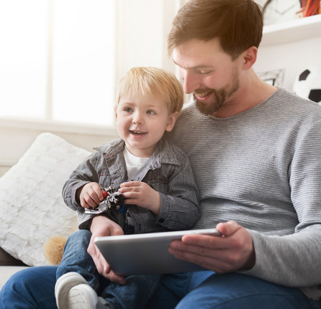 Happy father and little son playing on digital tablet sitting on sofa at homeの写真素材