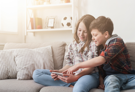 Two kids playing or watching video on smartphone while sitting on sofa at home.の写真素材
