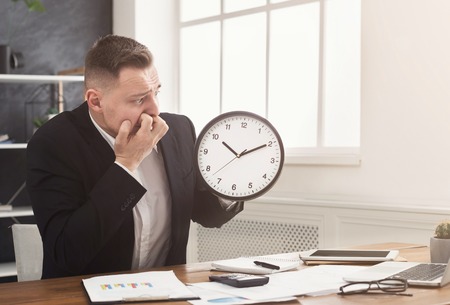 Worried businessman holding clock and looking at it. biting his fingers. Deadline and time limit concept, copy spaceの写真素材