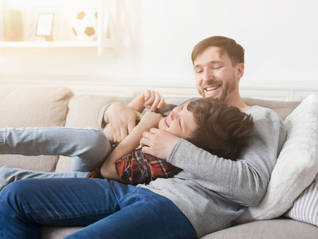 Loving father having fun and tickling his son on sofa at homeの写真素材