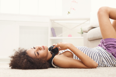 Smiling african-american girl dreaming, listening to music in headphones, lying on floor, relaxing at home, copy spaceの写真素材