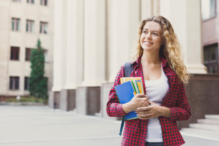 Beautiful smiling female student holding notebooks in hands while standing in front of the university campus. Education concept, copy spaceの写真素材