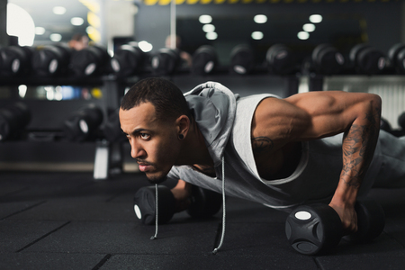Young african-american man workout in fitness club. Black guy making plank or push ups exercise on dumbbells, training indoors, copy spaceの写真素材