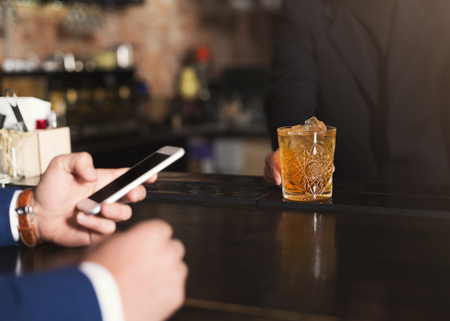 Bartender giving glass of whiskey to customer in bar, businessman using mobile phone, copy spaceの写真素材