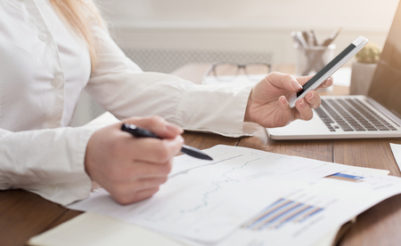 Young secretary using smartphone and reading documents at workplace in officeの写真素材