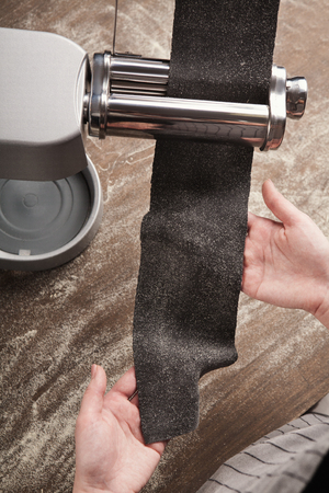 Rolling raw black dough with pasta machine, unrecognizable woman preparing homemade unusual colored fettuccine with cuttlefish ink, copy spaceの写真素材