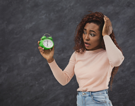 Upset african-american woman looks in shock at alarm clock over grey background