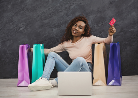 Young african woman shopping online at home. Girl sitting between colourful shopping bagsの写真素材