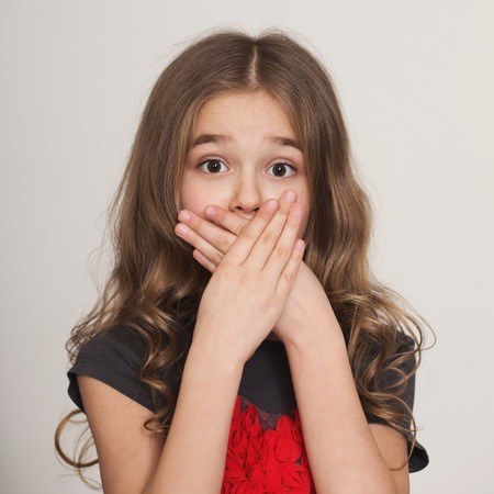 Shocked little girl covering her mouth by hands, looking at camera with wide-opened eyes