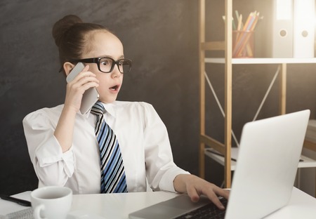 Shocking news. Little surprised girl using laptop and talking on phone in office, copy spaceの写真素材