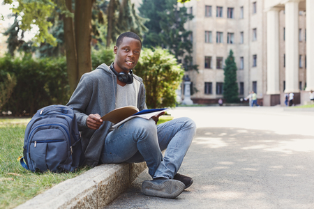 Happy african-american student sitting with notebook in university campus. Man studying outdoors with copybook. Education, inspiration and remote working concept.の写真素材