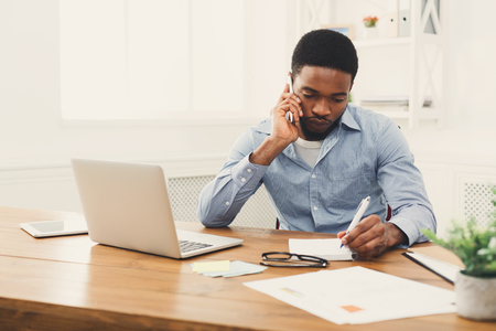 Young black businessman talking on mobile phone and making notes while working on laptop in modern white office, copy spaceの写真素材