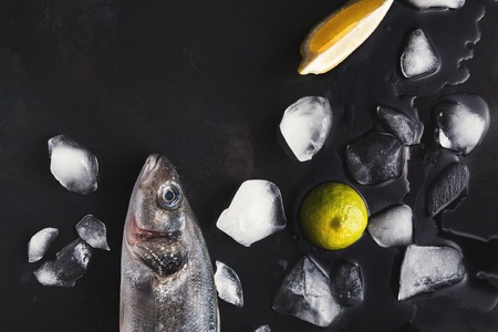 Raw sea bass head with ice and lemon on black background. Minimalistic mockup for seafood restaurant or fish market. Top view, copy spaceの写真素材