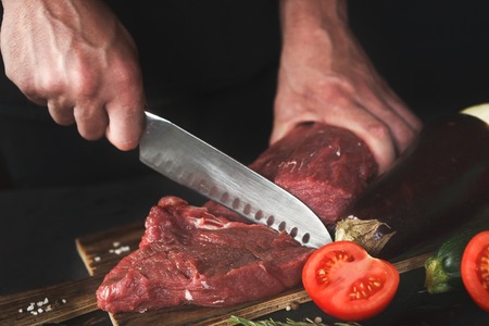 Man slicing filet mignon on wooden board at restaurant kitchen. Chef preparing fresh meat for cooking. Modern cuisine backgroung with herbs and vegetables, copy spaceの写真素材