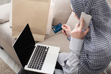 Arab woman making online purchase on laptop, ordering on smartphone, back viewの写真素材