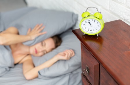 Young woman covering ears with pillow in bed while hear alarm clock ringingの写真素材