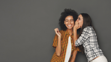Young woman whispering to her friend against grey background, copy space