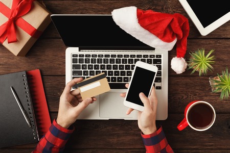 Man shopping online with credit card and smartphone with blank screen. Top view on human hands, keyboard, coffee, Santa hat, Christmas gift on wooden table background. New Year, holiday conceptの写真素材