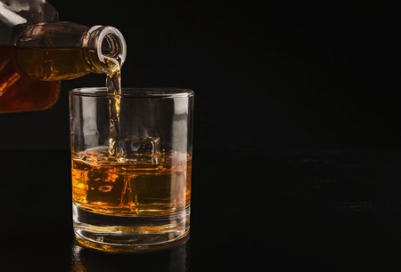 Pouring whiskey from bottle into glass with ice cubes on black background, copy spaceの写真素材
