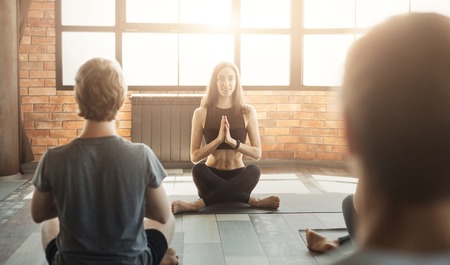 Group of young sporty people practicing yoga with instructor, sitting in Padmasana, making breathing exercise with folded hands in namaste, copy spaceの写真素材
