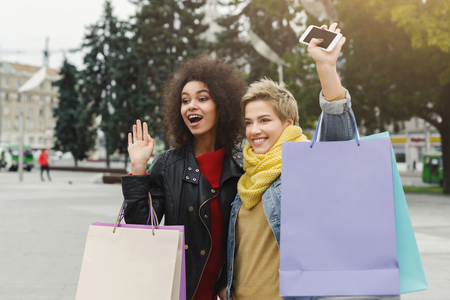 Happy girls with shopping bags greeting with someone. Female friends in bright warm casual clothes having a city walk. Friendship and leisure conceptの写真素材