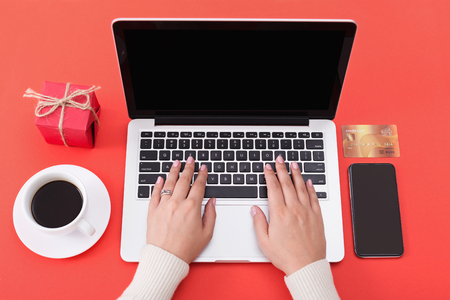 Woman buying christmas gifts online on laptop. Smartphone, credit card and gift box on red backgroundの写真素材