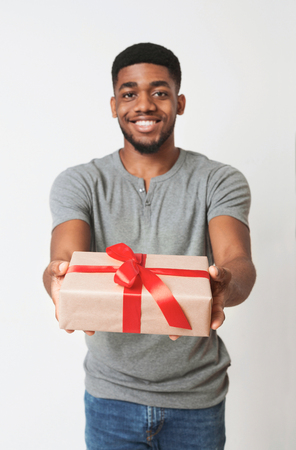 Theme holidays. Cheerful african-american man offering exclusive gift box wrapped with red ribbon and bow to cameraの写真素材