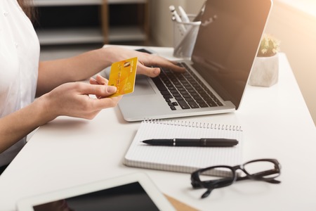 Unrecognizable woman at home shopping online on laptop, holding credit card, sitting at table in...