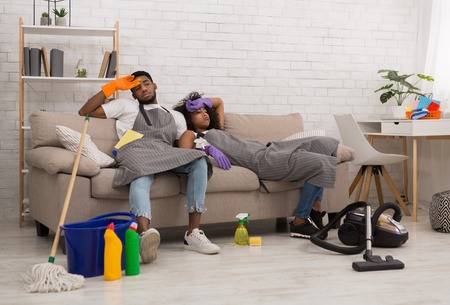 Tired young african-american couple is wiping foreheads while sitting on sofa after cleaning apartment, copy spaceの写真素材