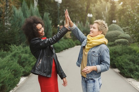 Outdoors portrait of two female multiethnic friends. Girls in casual warm outfits having a walk at park in cold season, having fun and giving high five.の写真素材