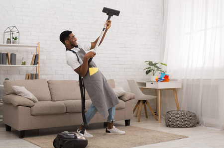 Happy african-american guy playing on vacuum cleaner like on guitar, having fun during house cleaningの写真素材