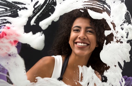 Young african-american housewife enjoying cleaning window glass, wiping out dense foam using spongeの写真素材