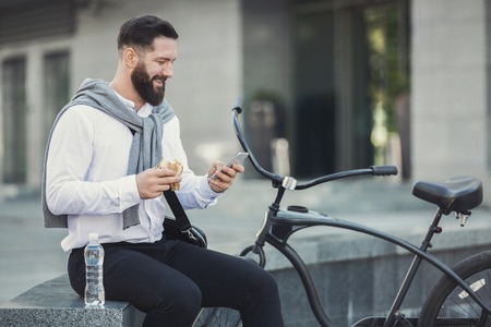 Bearded businessman went out for lunch on the street, eating burger and using smartphone, copy spaceの写真素材