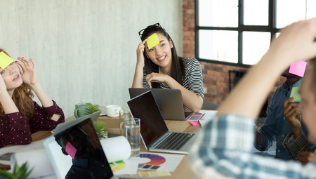 Young coworkers enjoying free time, raising togetherness and team spirit while sitting in office and playing game with paper stickers on foreheadの写真素材