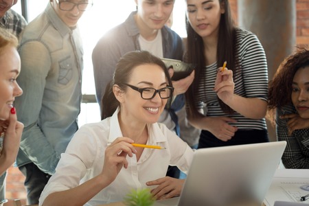Satisfied boss with her team. Smiling millennial professional people looking at laptop together, discussing projectの写真素材